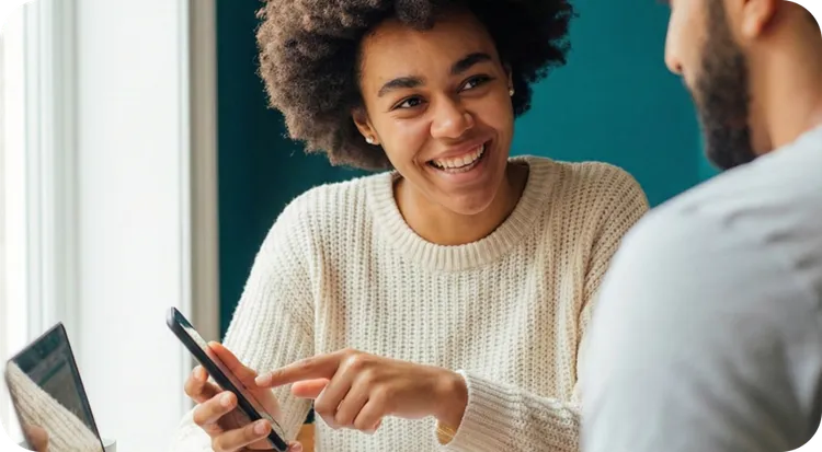 Young woman with curly hair and a white sweater smiling broadly and pointing at her smartphone screen while interacting with a coworker or client.