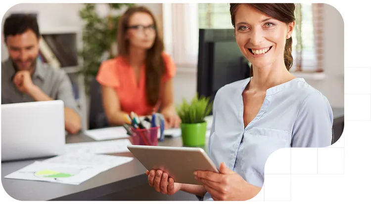 Profesional de servicios empresariales sonriendo y sosteniendo una tableta digital en una oficina moderna, mientras su equipo colabora al fondo en tareas de consultoría y análisis.