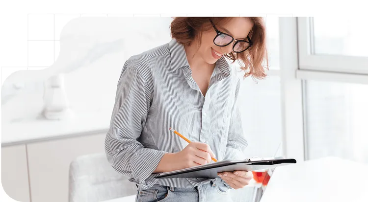 Mujer con gafas completando un formulario relacionado con firma electrónica reconocida en un portapapeles, en un espacio de trabajo luminoso junto a una ventana.